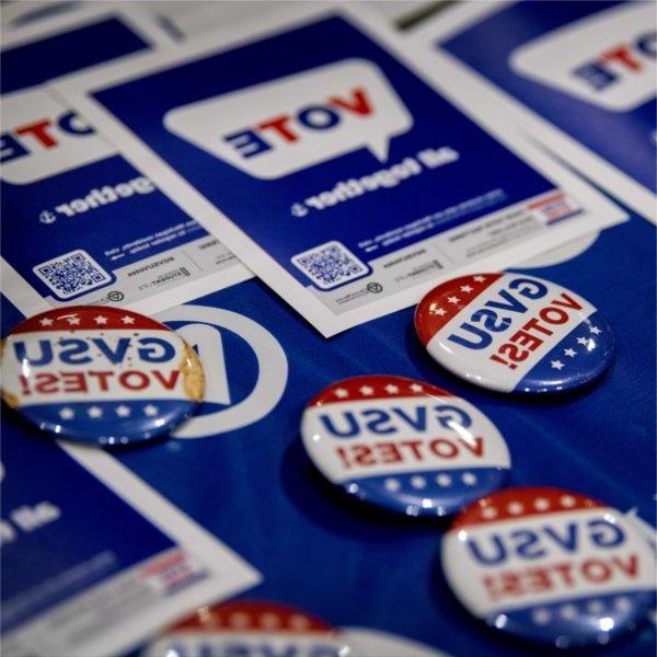 A collection of buttons that read �GVSU Votes!� in blue and red text sit on a blue Grand Valley tablecloth. Behind them are flyers with a QR code that say �Vote! All together�.