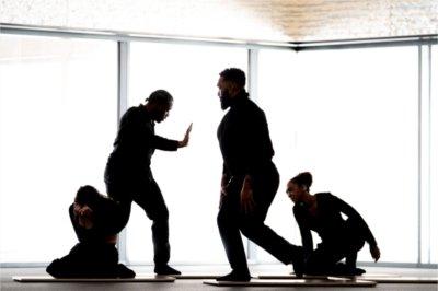 Dancers in all black perform modern movement in front of windows.